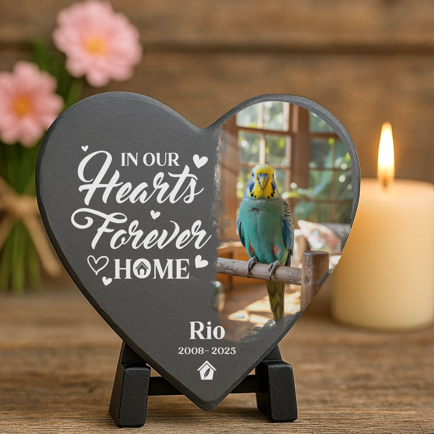 Heart-shaped bird memorial plaque on a stand, with candle and flowers in the background, displaying photo of parakeet Rio and touching remembrance quote.