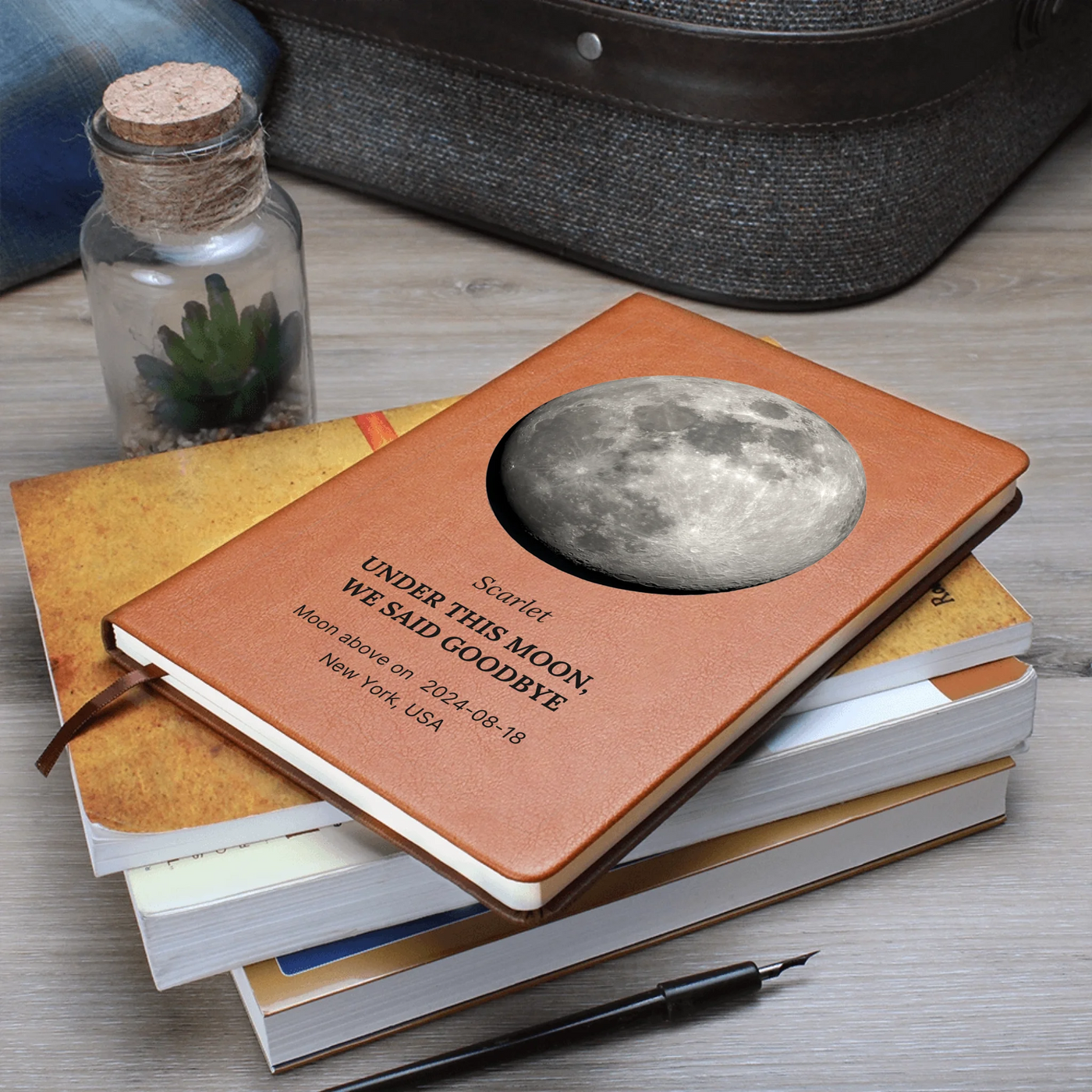 Moon-phase vegan-leather journal stacked with books and pen — custom date, location, and name printed on cover
