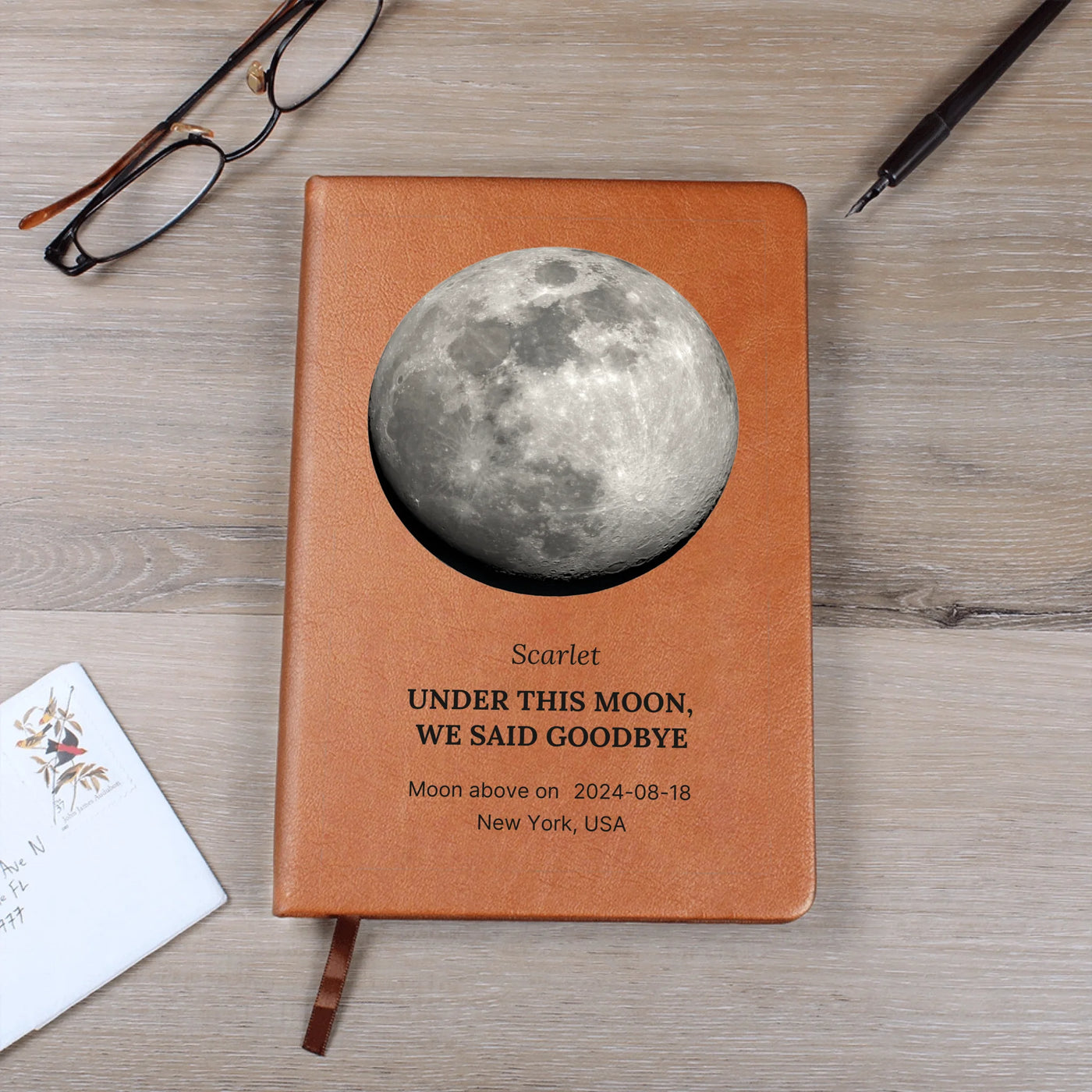 Flat lay: personalized moon-phase journal with gift tag — saddle vegan leather, custom name, date, and location