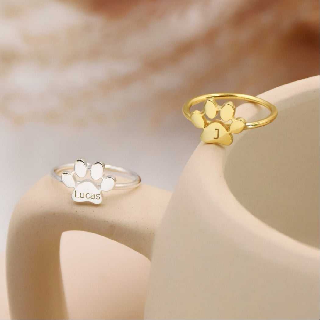 Two paw-shaped rings, one gold and one silver, on a ceramic surface with a blurred natural background.